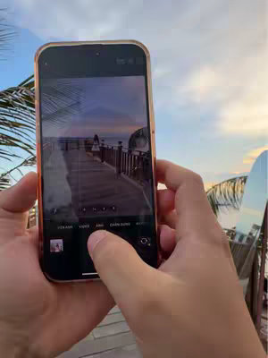 A person is holding an iPhone, capturing a photo of a wooden pier extending into the ocean. The phone's screen displays the sunset, which is partially obscured by the person's hands. The overall composition suggests a scenic moment being captured on a phone.