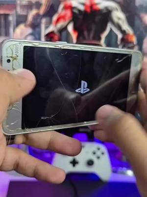 A close-up shot of a cracked smartphone held by two hands, displaying the Playstation logo on a black screen. Behind the phone, a blurry background features parts of an image, including the head and shoulders of Kratos from God of War. A white game controller is visible in the background