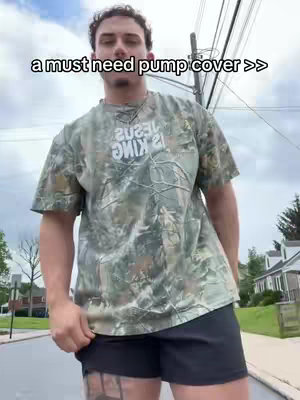 A man in a camouflage shirt and black shorts stands on a sidewalk, with a street and houses in the background. Text overlayed on the image reads, "a must need pump cover >>".