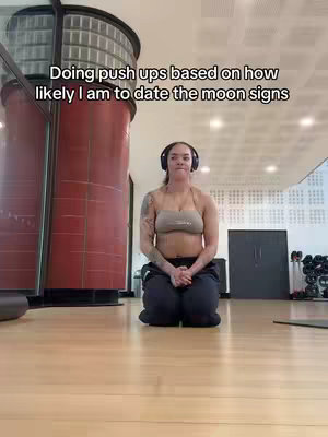 A woman kneels on a wooden gym floor, wearing a sports bra and sweatpants. She has tattoos on her arm and is wearing headphones; text overlaid states, "Doing pushups based on how likely I am to date the moon signs."