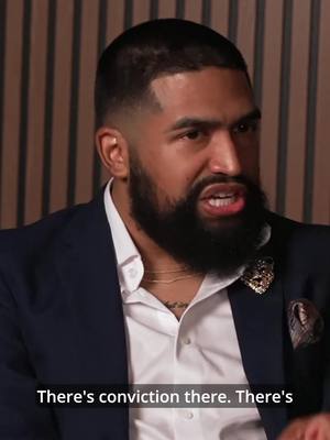 A medium shot features a man with dark skin and a neatly trimmed beard, wearing a dark blue blazer over a crisp white shirt. He has a serious expression, with his mouth open as if speaking with conviction. The background is a textured wall with vertical stripes.