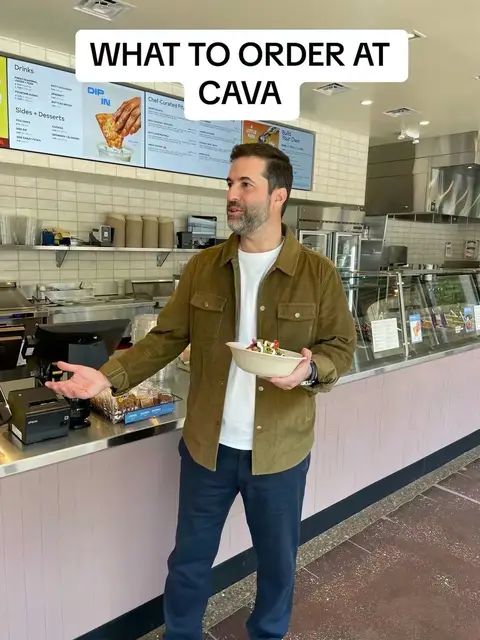 A man stands in a CAVA restaurant holding a bowl of food. He is gesturing with his left hand while looking at the camera. The background shows the restaurant's counter and menu.