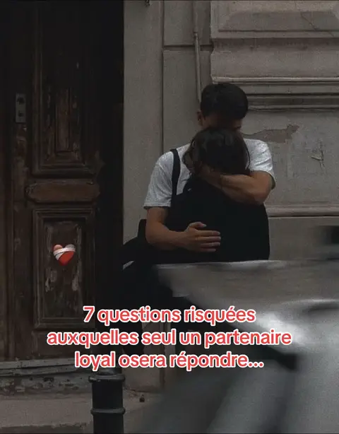 A photograph depicts an embrace between two people, likely a couple, in a dimly lit outdoor setting. The primary subject seems to be a man hugging a person from the back, whose face is obscured. The background includes a dark doorway and a section of a concrete wall. The image has a warm, slightly faded color palette. Text overlay reads '7 questions risquées auxquelles seul un partenaire loyal osera répondre...' 