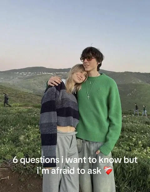 A young couple embraces outdoors, set against a mountain backdrop. The woman rests her head on the man's shoulder, both facing forward. Text overlays the bottom of the image.
