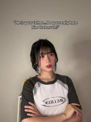 The cover image shows a young woman with black hair and bangs, positioned in front of a light-colored background. Overlayed text in the upper-center reads, "He is your father... Do you really hate him that much!" The woman is wearing a two-tone t-shirt with short sleeves, black on the sleeves and shoulders, and white with a logo in the center. Her arms are crossed, and she is looking slightly off-camera with a neutral expression.