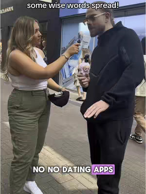 Two people are standing on what appears to be a city street. A woman on the left is holding a microphone and speaking to a man with a beard and glasses on the right. There is text overlayed on the image. The woman wears a white tank top and green pants. The man is dressed in all black and holding a microphone and something pink.