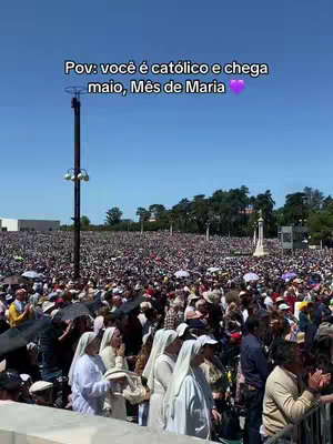 A wide shot of a large crowd of people gathered outdoors with a clear blue sky overhead. The crowd appears dense, with a mix of individuals, some wearing religious attire. A tall pole with lights stands in the center-left, and a white building topped with a statue is visible in the background. The image is bright and filled with natural light.