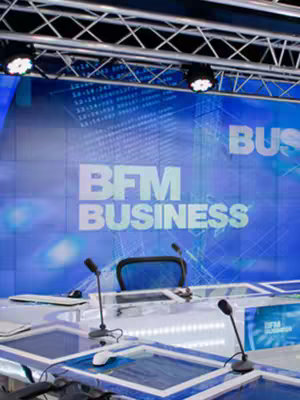 The image captures the interior of the BFM Business television studio. The background features a large screen displaying the channel's logo and branding. In the foreground, there's a desk with microphones and a computer setup, seemingly prepared for a broadcast. The overall composition is professional, with a focus on the studio environment.