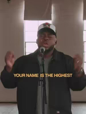 A man stands in front of a microphone, arms raised, with a plain background featuring windows. Text overlayed at the bottom reads 'YOUR NAME IS THE HIGHEST'.