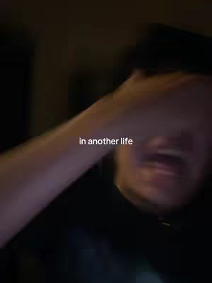 A blurred selfie-style video shows a person's face and arm. The person is holding their arm up, partially covering their face. The lighting is dim, and the background is indistinct. Text overlayed onto the image reads 'in another life'.