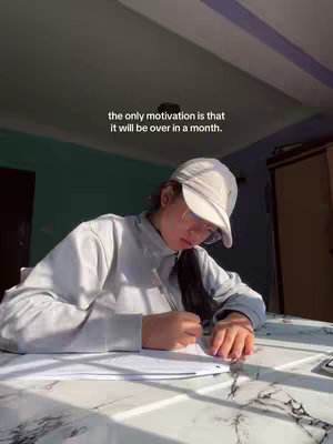 A person wearing a cap and glasses is sitting at a table, writing on a piece of paper. The person is the focal point, with the background showing a room. Text overlayed on the image reads 'the only motivation is that it will be over in a month.'