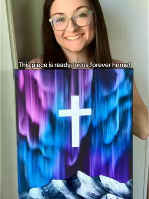 A woman with glasses smiles, holding up a painting featuring a cross and a landscape of a colorful sky and mountains.