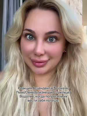 A close-up shot of a woman with blonde wavy hair and blue eyes. She is centered in the frame, looking directly at the camera with a slight smile. The background is blurred, suggesting an indoor setting with natural light.