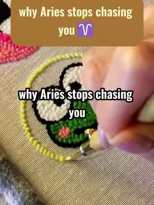 The image focuses on a hand using a craft punch needle tool to create an image on a piece of fabric. The element being created is a green frog with white eyes, and it is enclosed in a yellow circle
