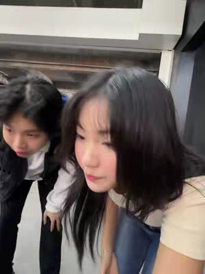 Two young women are leaning downward with their heads close together, their faces visible. The background is a blurry interior setting, and there is a white surface on the right. The woman to the left has short dark hair. The woman on the right has long dark hair with a fair complexion.