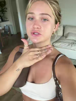 A woman with blonde hair is looking at the camera from a medium angle, slightly tilting her head as she holds her hand up to her neck. She's wearing a white tank top, and a brown purse strap is visible on her right shoulder. The background is a softly lit room with a bed and some other furnishings visible.