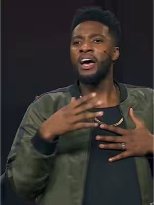 A man of African descent is speaking with animated hand gestures. He is standing; the background is dark, likely a stage. He's wearing a black shirt under an olive-green bomber jacket and a subtle gold necklace.