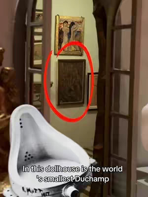 The cover image showcases a dollhouse interior with detailed architectural elements. The focal point is a red circle drawn on the image highlighting a framed painting on the white wall. Adjacent to the wall is a white toilet with black details The frame is constructed with a combination of warm brown wooden features. The image overall has a warm-toned and realistic aesthetic, capturing a sense of depth and artistic focus on a tiny dollhouse.