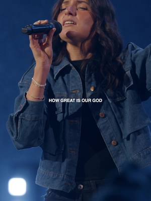 Image depicts a mid-shot of a woman singing into a microphone. She wears a denim jacket and has dark hair. Overlaid text reads, 'HOW GREAT IS OUR GOD'. The background is blurred with dark blue and dim lighting.
