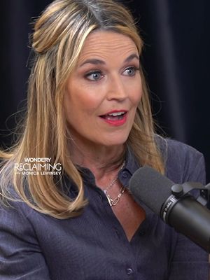 A medium shot of a woman with blonde hair styled partially up. She's looking slightly off-camera with a surprised expression and red lipstick. A microphone is in the background, suggesting an interview or podcast setting with a dark background.