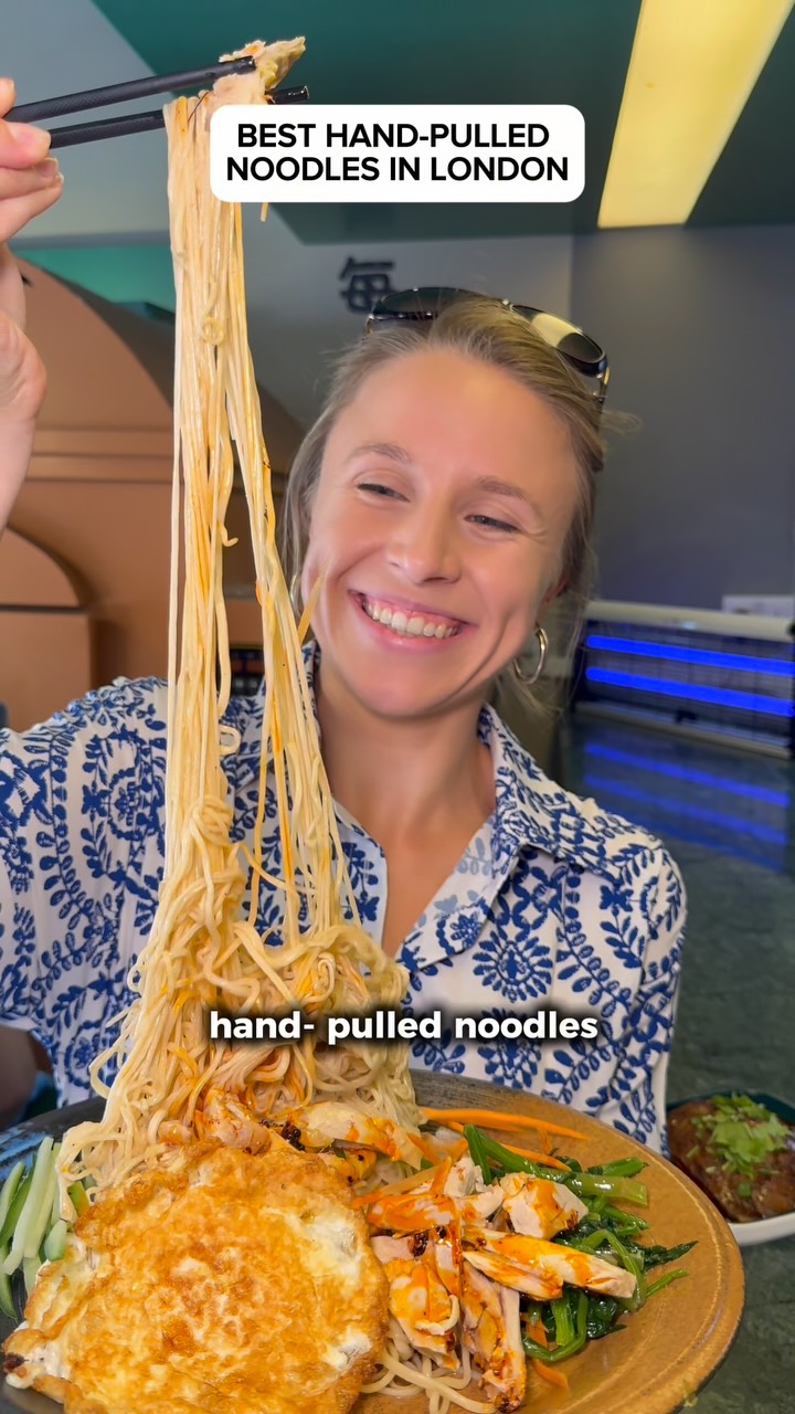 A woman with blonde hair and sunglasses on her head is smiling while holding a pair of chopsticks with noodles held high. Under the chopsticks are long noodles hanging down over a plate of food. The plate includes a fried egg, chicken, sauce, chopped vegetables and noodles. In the background is the interior of a restaurant setting with another plate of food.