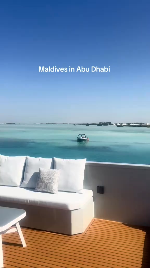 The cover image features a serene outdoor scene.  A light-colored bench with white cushions and a patterned pillow sits on a wooden deck.  The scene overlooks a turquoise body of water dotted with small green islands.   A small boat is visible, gliding across the water. A clear blue sky serves as the backdrop.  The image evokes a sense of luxury and tranquility.