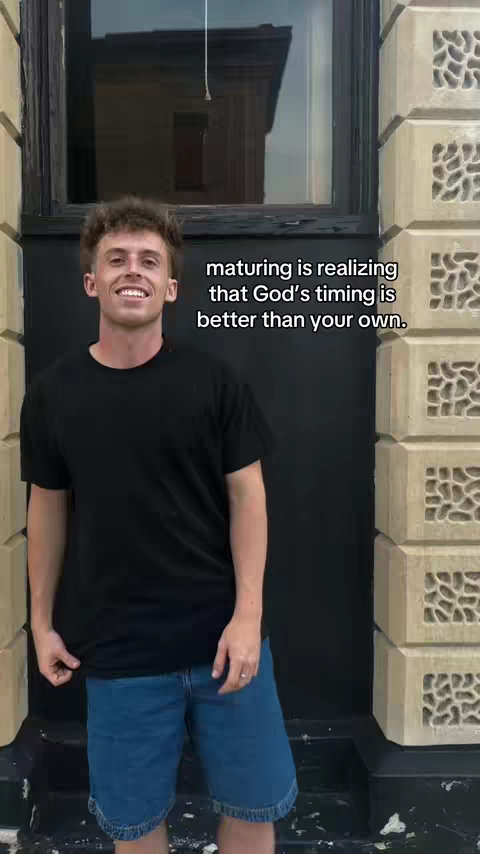 A young man stands angled slightly to the left, smiling at the camera. He wears a black t-shirt and blue jean shorts, stands against a black backdrop with light-colored building elements to each side. Text in the upper right corner reads "maturing is realizing that God's timing is better than your own."