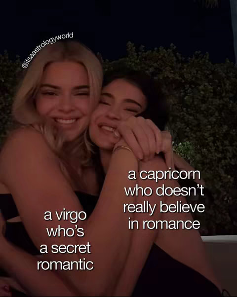 Two young women are embracing in a close-up shot. They are smiling and appear to be close friends. Text overlays are superimposed on the image, describing the two as a virgo who's a secret romantic, and a capricorn who doesn't really believe in romance.