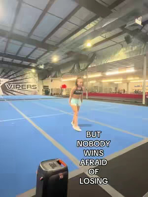 A young girl is walking towards the viewer in a gymnasium. She's wearing sportswear and appears to be on a blue mat. Text overlay says, 'But nobody wins afraid of losing'. There is a visible speaker in the lower left corner.