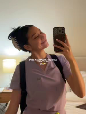 A selfie-style photo of a young woman in light purple scrubs, holding a phone up to her face with her right hand and looking at it.  She has dark hair pulled back with a claw clip.  The background is a softly lit room.