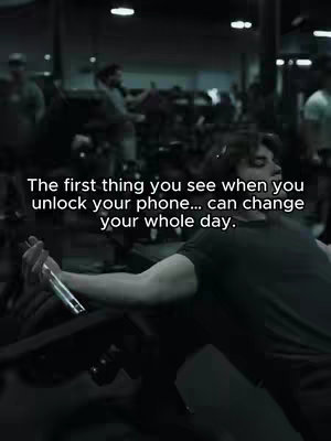A grayscale image of a man working out in a gym. He is wearing a dark t-shirt, and visible is his arm, holding a dumbbell. The gym is dimly lit with several other people in the background, some appearing to use gym equipment. Text overlayed on the image states, "The first thing you see when you unlock your phone... can change your whole day."