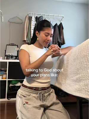 A young woman kneels in front of a bed, hands clasped in a prayerful posture. The room has a pale pastel color scheme, with a rack of clothing visible behind her. The image composition includes text overlay that states 'talking to God about your plans like:'
