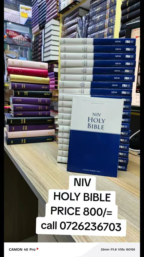 A shop display featuring stacks of Bibles. The foreground highlights a row of stacked books with blue and white covers, one of which is slightly open. Behind, other varieties of books are visible. The lighting is bright, common to shop settings.