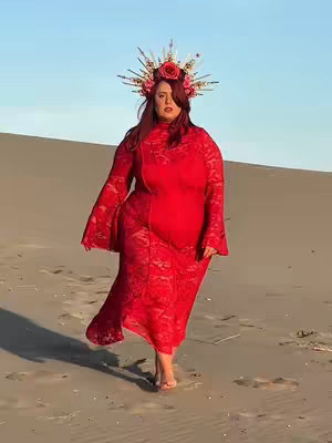 A woman in a long red dress and elaborate headpiece walks on a sandy beach, with a bright blue sky in the background. The dress has a lace texture. The woman's hair is red.