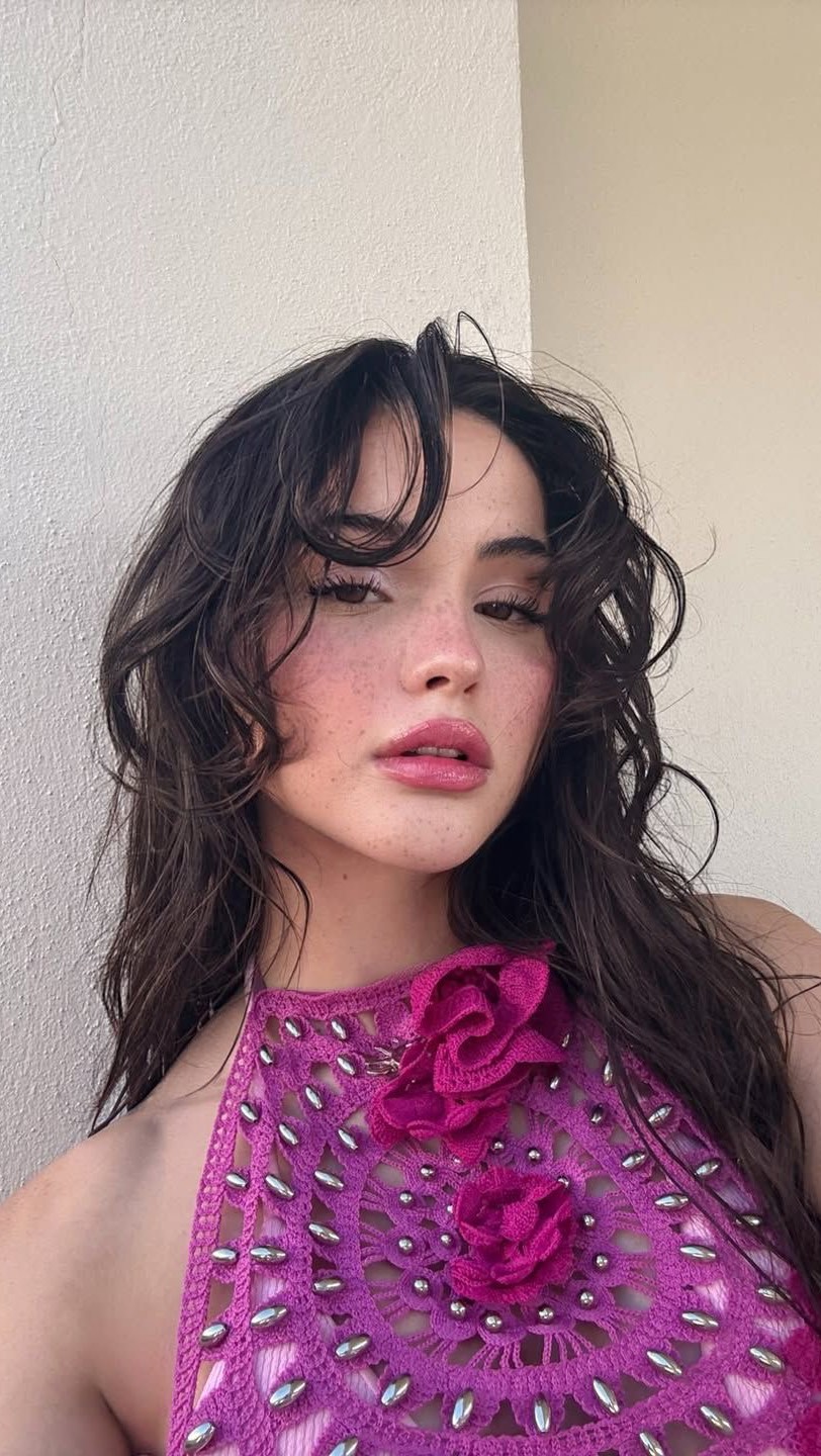A close-up selfie of a woman with long, dark, wavy hair and a pink halter top. She has freckles on her face, pink lips, and is looking directly at the camera. The wall behind her is plain and beige.