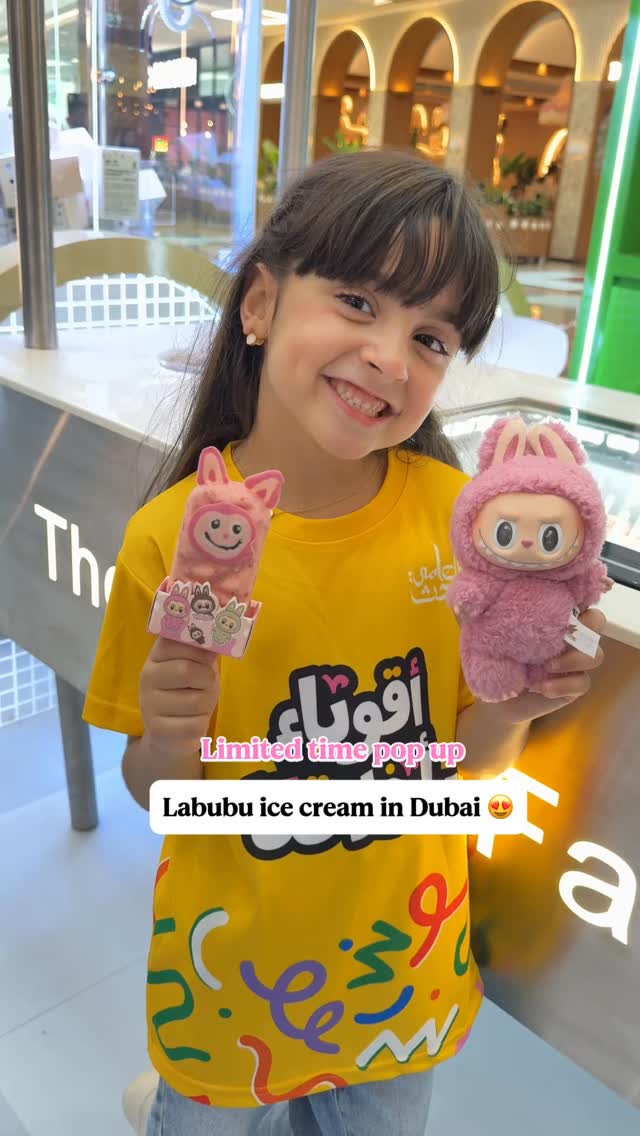 A young girl with long brown hair is smiling, holding a pink Labubu doll and a Labubu ice cream bar. She is wearing a yellow shirt with printed designs, in a bright indoor setting.