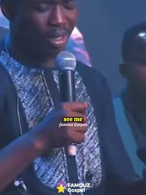 A medium shot of a Black man singing into a microphone, with overlaid text. The man is wearing a black patterned shirt. The background is dark and out of focus. There is added text on the image.
