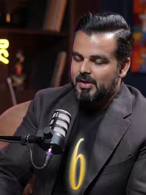 The image shows a man, appearing to be in a podcast studio. He is wearing a black t-shirt with a stylized gold number 6. He has a beard, and is facing forward, holding a microphone. Behind him, there are bookshelves.