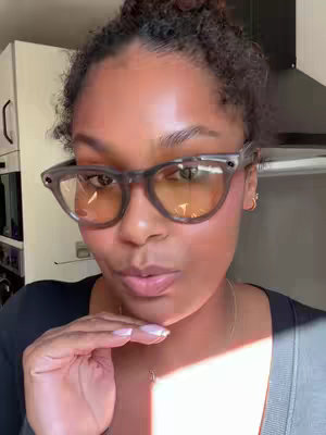A woman with dark curly hair is shown in a close-up shot, wearing clear-framed glasses with yellow-tinted lenses. She holds her hand up, fingers near her lips, giving a pensive look.