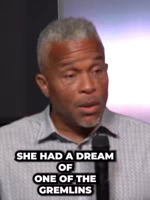 The cover image shows a close-up of a middle-aged Black man with short, graying hair. He is wearing a light gray, button-down collared shirt and a microphone. The man appears to be speaking. Overlayed on the image, there is text in white font with a black outline that reads "SHE HAD A DREAM OF ONE OF THE GREMLINS."