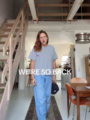 A woman with auburn hair stands in a brightly lit room. She wears a light blue t-shirt, light blue jeans, and has a black hat in her hand. The text "WE'RE SO BACK" is overlaid on the image in white.