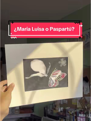 The cover image shows an artwork held up by a hand. The artwork itself is a drawing of two butterfly-like figures on a dark gray background, framed by a white mat. The background behind the hand appears to be a room with additional items visible to the side.