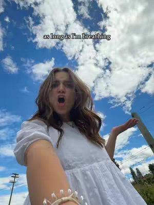 A young woman in a white dress is seen from a low angle, taking a selfie with a blue sky and fluffy clouds in the background. Her mouth is open and her expression is one of surprise or excitement. Text overlay reads: as long as I'm breathing