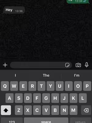 Screenshot of a mobile phone displaying a messaging app interface. The background is a dark, patterned design. The bottom half of the screen shows a keyboard with white keys and gray accents, including the 'space' and 'return' keys. Above the keyboard is a text input field, and above that, a chat bubble that says 'Hey,' with a timestamp '13:36.' The top right shows a charging and signal info. The overall tone suggests communication or a tutorial on texting.