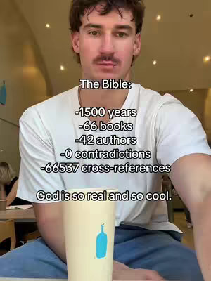 A man is centered in the frame, holding a coffee cup. Text overlayed on the image lists facts about the Bible. The man is fair-skinned with short brown hair, a mustache, and a white t-shirt. The background is a bright, open space, likely an indoor cafe.