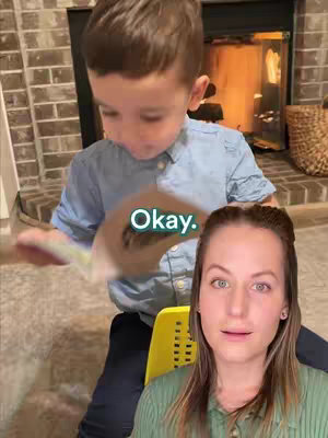 A cover image shows a young boy holding a book, appearing to be looking at the content of the book. His eyes are focused on the pages, and he is positioned in the upper center of the frame. A woman, likely the person filming, is in the foreground, facing the viewer with a concerned expression. Behind the boy, a brick fireplace and a glimpse of a fire are visible. A text overlay saying "Okay" is on the book. Additional element is a yellow chair.