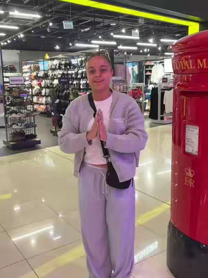 A woman stands in a well-lit retail store, facing the camera with hands together in a prayer-like gesture. She wears a light gray sweatsuit and a white top. A black crossbody bag hangs across her chest. A red postbox is visible on the right of the frame. The background is a store interior with shelves of merchandise.
