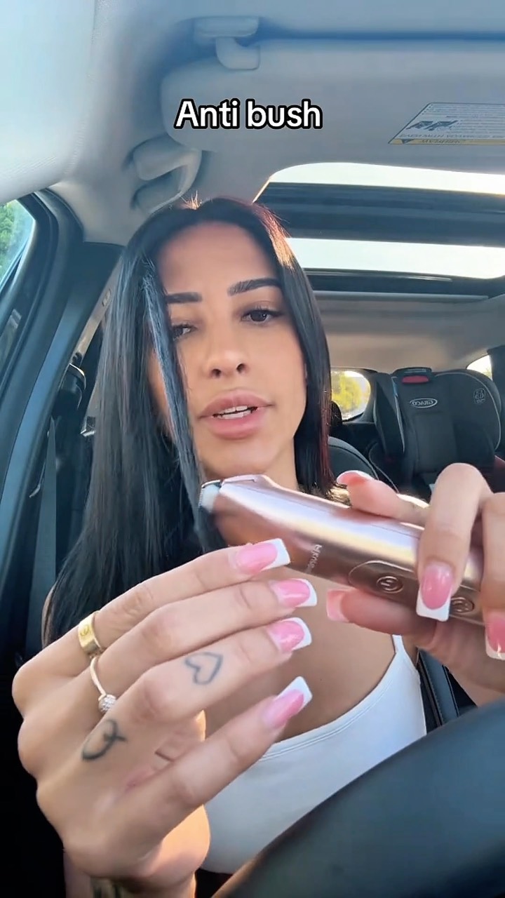 An attractive woman is shown in the driver's seat of a car, holding a gold and rose-gold electric razor. She is looking at the camera while holding the razor, her nails are painted a white and pink french tip. The text overlay on the image says "Anti bush".