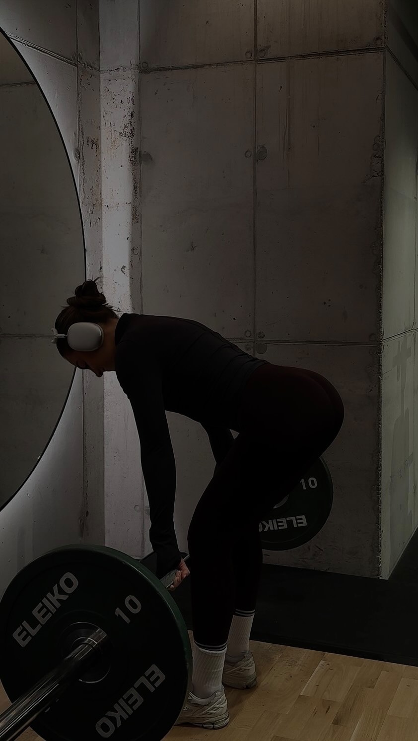 A woman in a gym is bent over, holding a barbell during what looks like deadlifts.  She wears a black long-sleeved top, black leggings, and white socks and sneakers.  She is wearing headphones, focused on the exercise. The background is a stark concrete wall and a circular mirror, gym equipment is in the foreground.