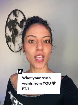 A woman with dark hair, pulled back, and wearing a black top, looks directly at the camera with her mouth open as if speaking. Behind her is a clock with a world map design and a plain wall. Overlayed on the image is a white box with the text, 'What your crush wants from YOU 🖤 PT.1'.